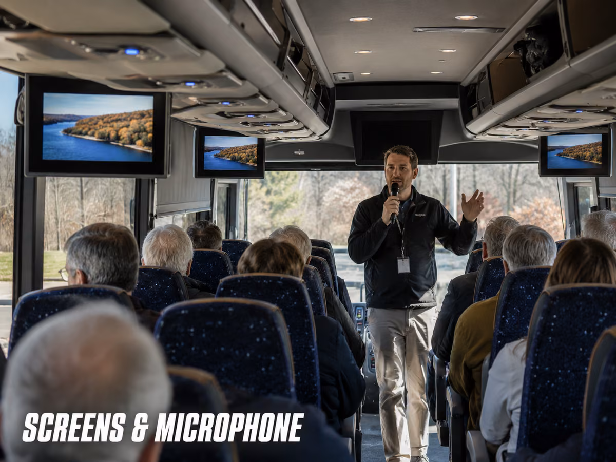 Passengers seated on a charter bus with overhead monitors visible down the aisle