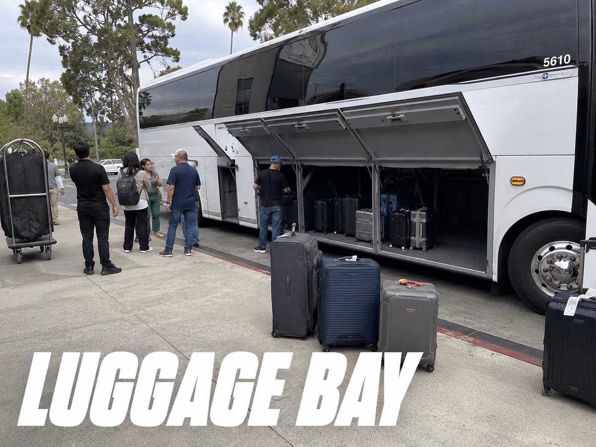 Passengers loading suitcases into the open undercarriage luggage bay of a motorcoach.