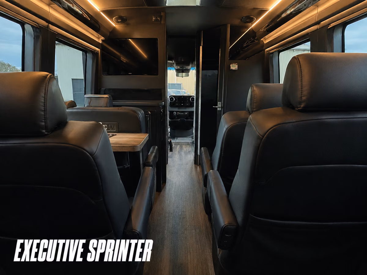 Interior of an executive Sprinter with individual forward-facing black leather captain's chairs and a small wood table.